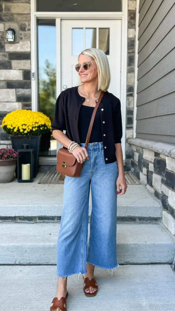 Blonde woman wearing high-waisted cropped wide-leg jeans with raw hems, a fitted black top, short-sleeve cardigan, and brown leather slide sandals for a chic summer outfit.