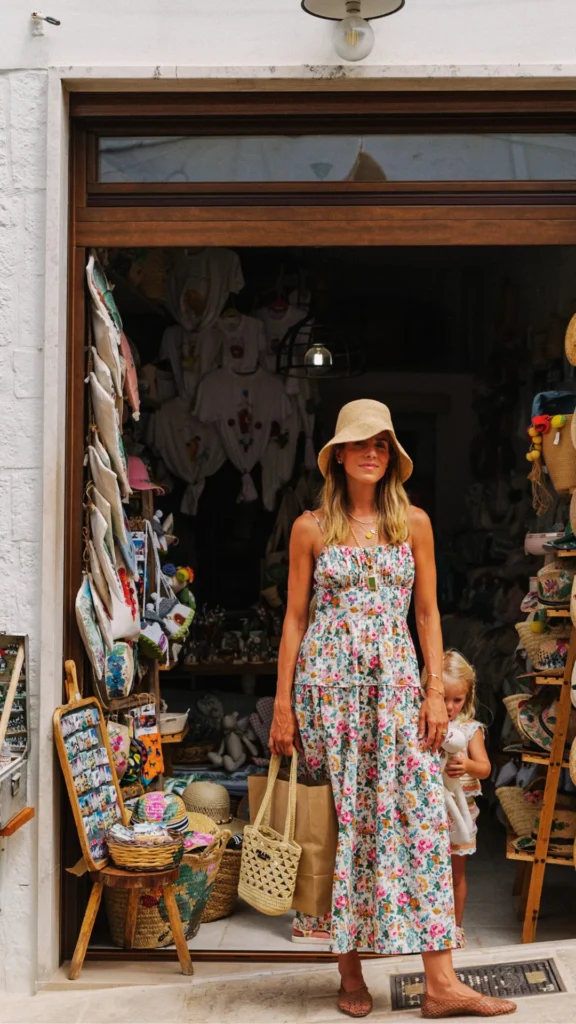 Woman wearing a floral print tiered summer midi dress, a straw bucket hat, woven flats, and carrying a woven straw tote bag outside a boutique store.