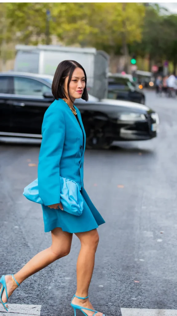 Woman with a sleek dark bob hairstyle wearing a vibrant monochromatic turquoise blue blazer, matching short skirt, sky blue pouch clutch, and bright blue strappy heel sandals.
