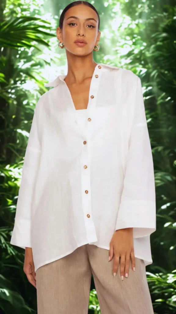 Woman wearing an oversized white linen button-down shirt layered unbuttoned over a white top, paired with beige linen trousers and gold hoop earrings for a breezy summer outfit.